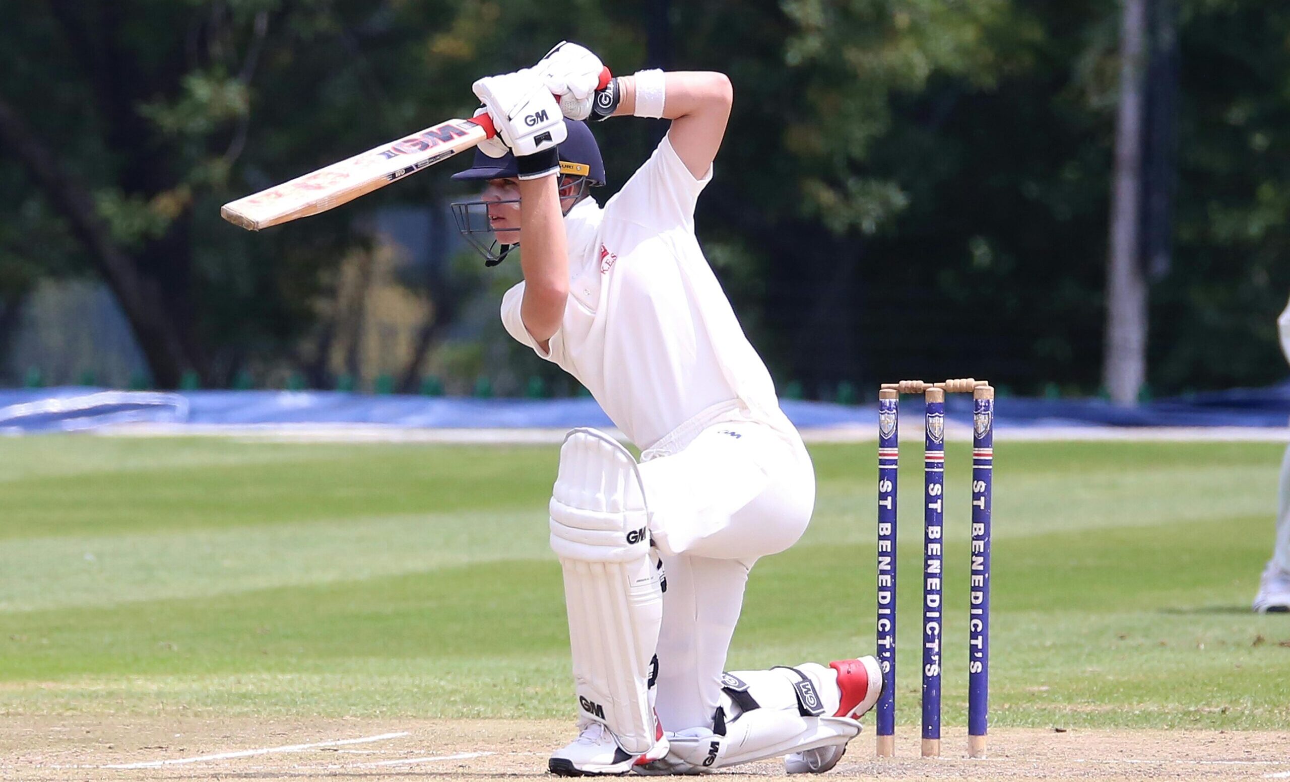 Cricketer in action performing a sweep shot on a sunny outdoor pitch.