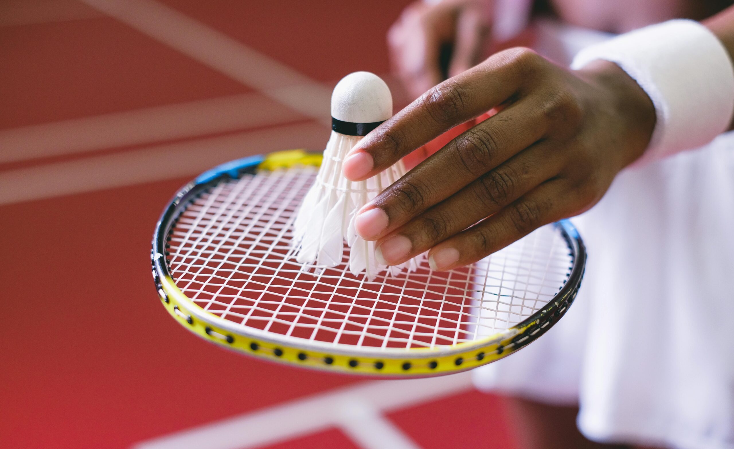 A detailed view of a hand holding a shuttlecock over a badminton racket, on an indoor court.